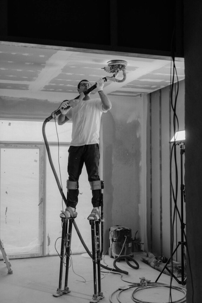 Worker using stilts and power sander on ceiling in construction site.